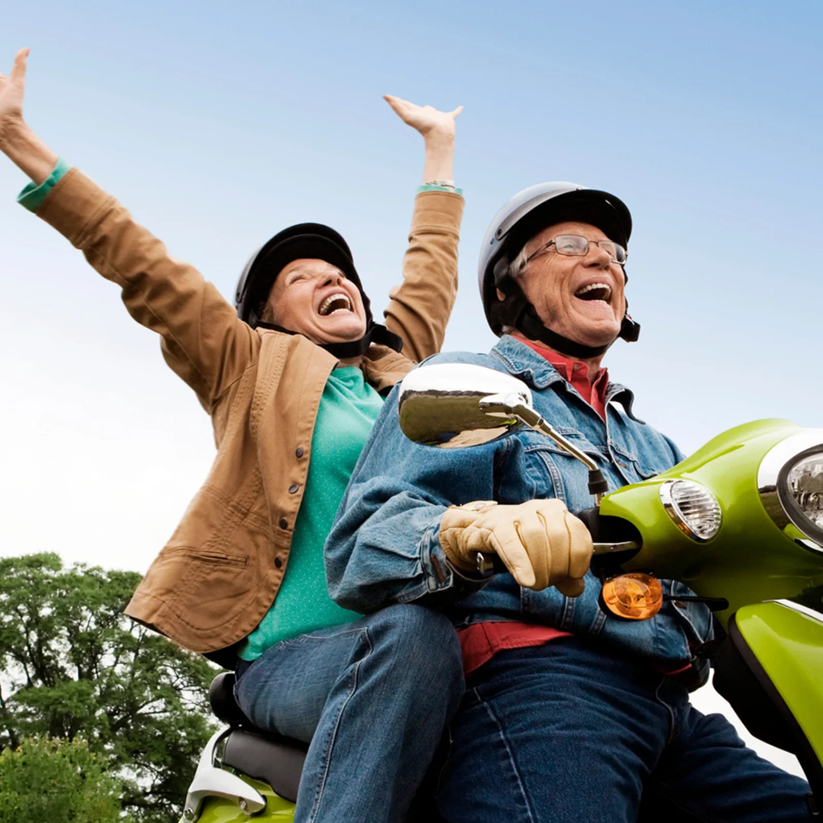 A retired couple riding a scooter with their arms raised, laughing